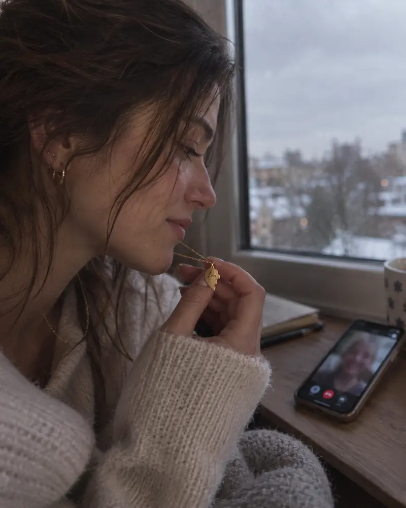 Une jeune femme tient son pendentif en souriant pendant un appel vidéo ; dehors, la neige tombe